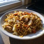 A close-up of a white plate filled with homemade Chicken Lo Mein noodles, chicken pieces, cabbage, and carrots.