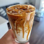 Close-up of a tall glass of Iced Caramel Swirl Coffee showing beautiful swirls of caramel and coffee mixing into the white milk base over ice.
