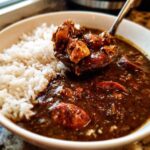 A spoonful of rich Justin Wilson Chicken and Sausage Gumbo, featuring chicken and sausage pieces, being lifted from a bowl next to fluffy white rice.