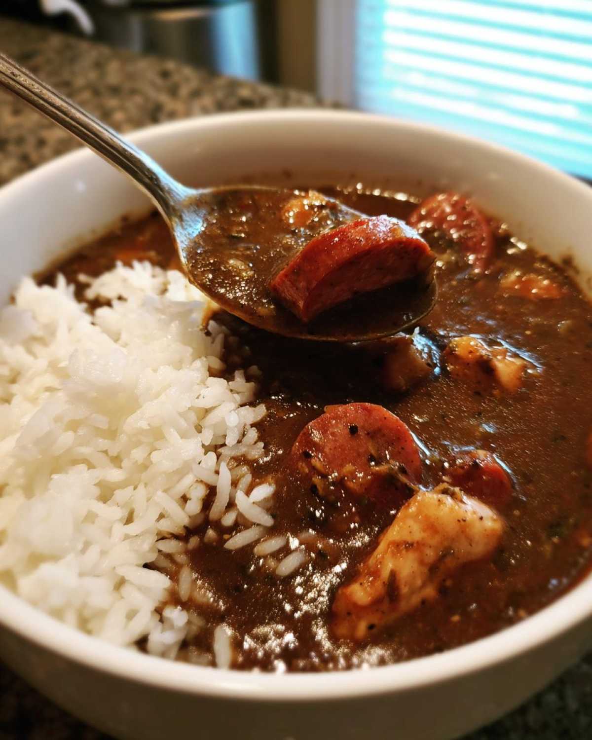 A spoonful of rich Justin Wilson Chicken and Sausage Gumbo, featuring sausage and chicken, being lifted from a bowl next to white rice.