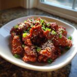 Close-up of crispy Korean BBQ Chicken pieces coated in a shiny red glaze, topped with sesame seeds and green onions.