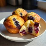 Four golden-brown Lemon Blueberry Muffins on a white plate, one is broken open showing juicy blueberries inside.