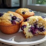 Three freshly baked Lemon Blueberry Muffins on a white plate, one broken open showing moist interior and bursting blueberries.