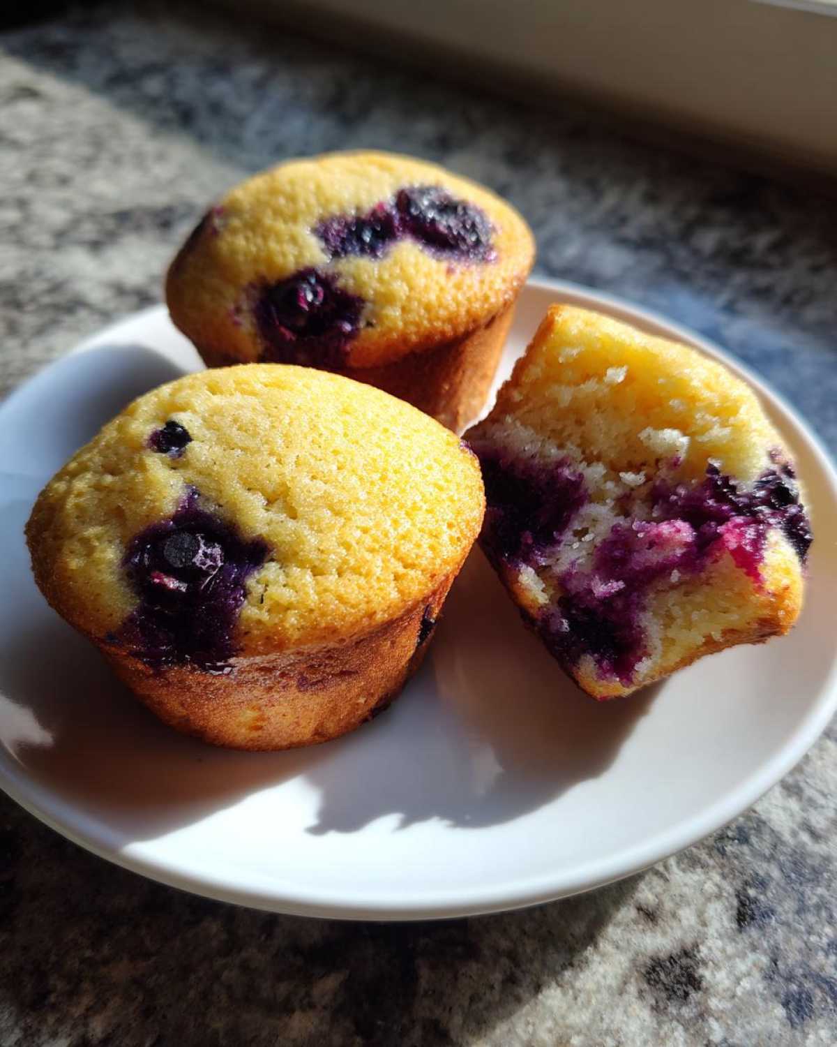 Three freshly baked Lemon Blueberry Muffins, one broken open to show the moist interior, sitting on a white plate.