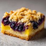 A close-up, macro shot of a single serving of Lemon Blueberry Streusel Bars showing the golden crust, bright lemon filling, and dark blueberries under a crumbly streusel topping.