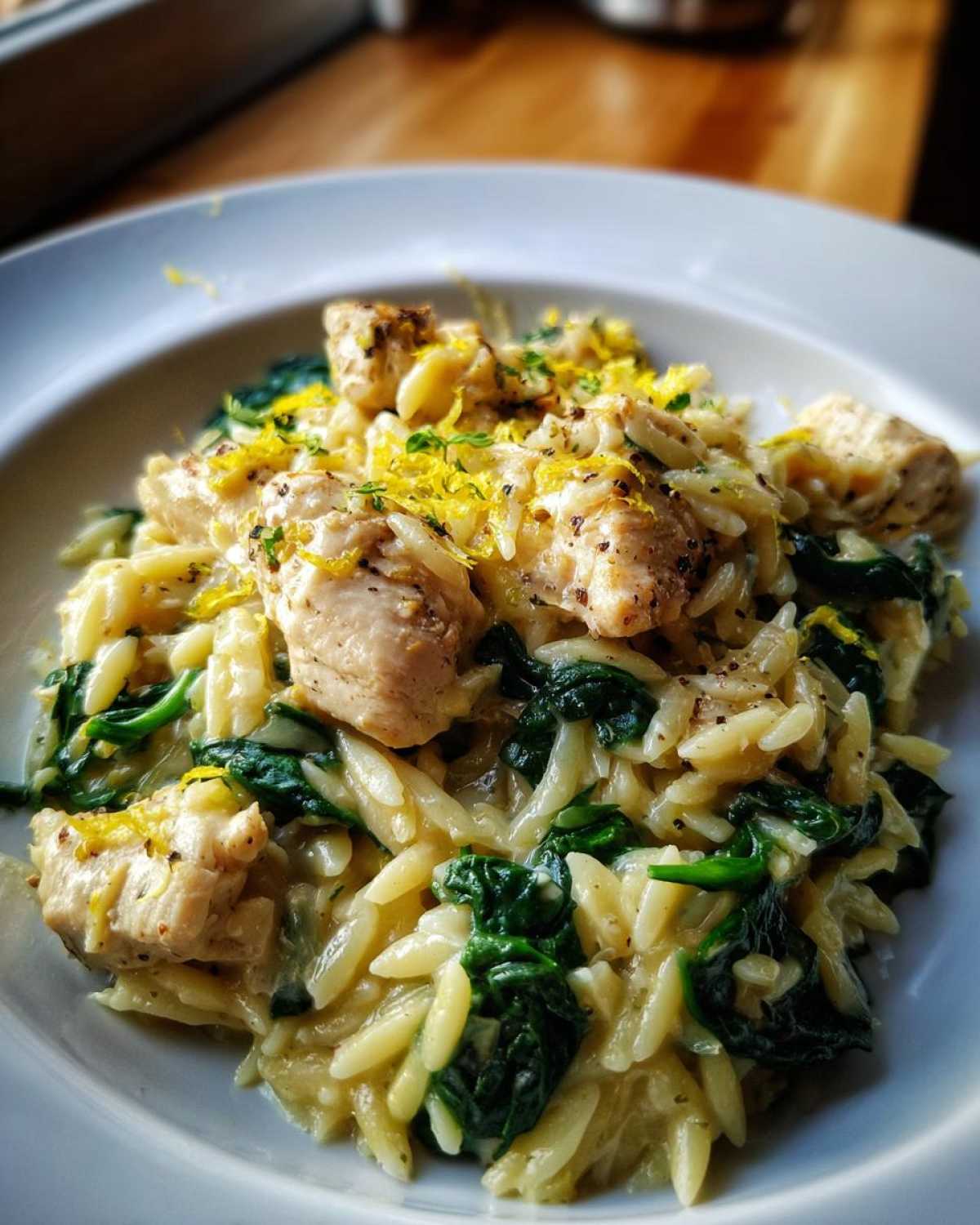 A close-up serving of creamy Lemon Chicken Orzo Spinach topped with bright yellow lemon zest and black pepper.