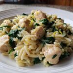 A close-up of a serving of Lemon Chicken Orzo Spinach on a white plate, featuring orzo pasta, chicken chunks, and wilted spinach.