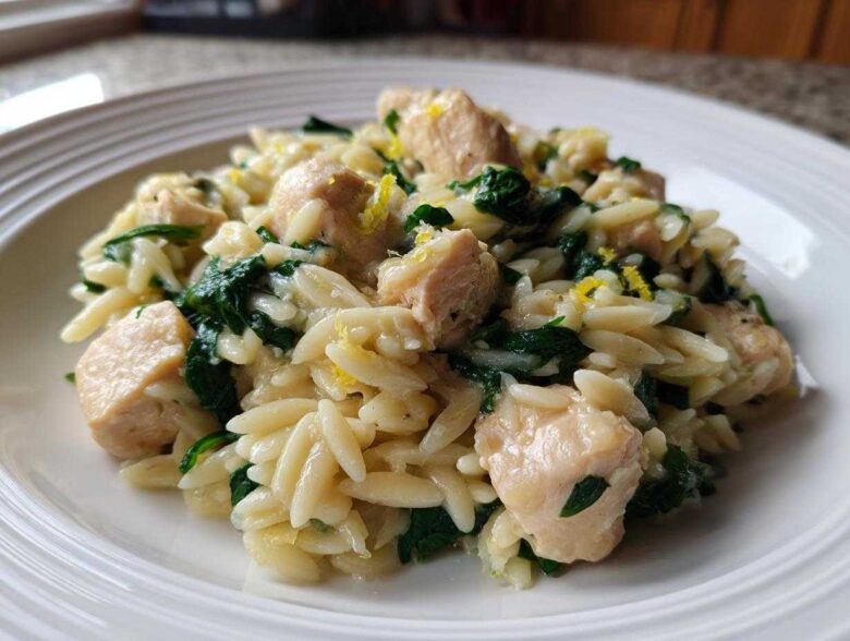 A close-up of a serving of Lemon Chicken Orzo Spinach on a white plate, featuring orzo pasta, chicken chunks, and wilted spinach.