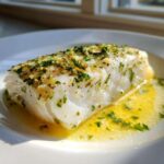 A close-up of a perfectly cooked white fish fillet, likely Lemon Garlic Butter Cod, resting in melted butter sauce and topped with herbs.