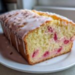 A loaf of moist Lemon Rhubarb Bread, sliced to show pink rhubarb pieces, topped with a white lemon glaze.