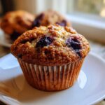 A close-up of a moist Banana Blueberry Muffin sitting on a small white plate, with more muffins blurred in the background.