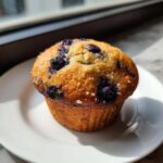 A close-up of a single, golden-brown Banana Blueberry Muffins sitting on a white plate near a window.