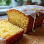 A close-up of moist Lemon Bread with thick white glaze dripping down the sides and lemon zest sprinkled on top.