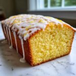 Close-up of a moist Lemon Loaf cake slice showing the crumb, topped with dripping white glaze and lemon zest.