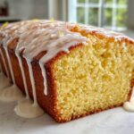 A close-up of a moist Lemon Loaf cake with thick white glaze dripping down the sides, garnished with zest.