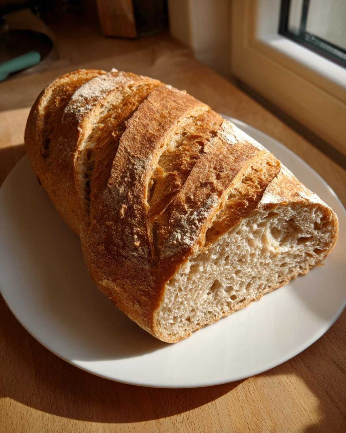 A freshly baked loaf of Quick and French Bread from This Pilgrim Life, sliced and resting on a white plate in sunlight.