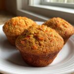 Close-up of three golden brown Quick & Cinnamon Carrot Muffins studded with visible shreds of orange carrot on a white plate.