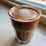 A close-up of a thick, dark brown Coffee Smoothie with a foamy top, served in a clear glass on a light surface near a window.