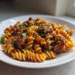 A close-up of fusilli pasta mixed with sausage pieces coated in a sticky honey garlic sauce, garnished with parsley.
