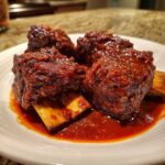 Close-up of four tender Crock Pot Short Ribs coated in a thick, glossy, dark red sauce, resting on a white plate.