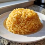 A mound of golden yellow Texas Roadhouse Seasoned Rice served on a white plate, brightly lit by sunlight.