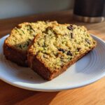Two thick slices of moist Zucchini Bread, showing shredded zucchini and dark fruit pieces, served on a white plate.